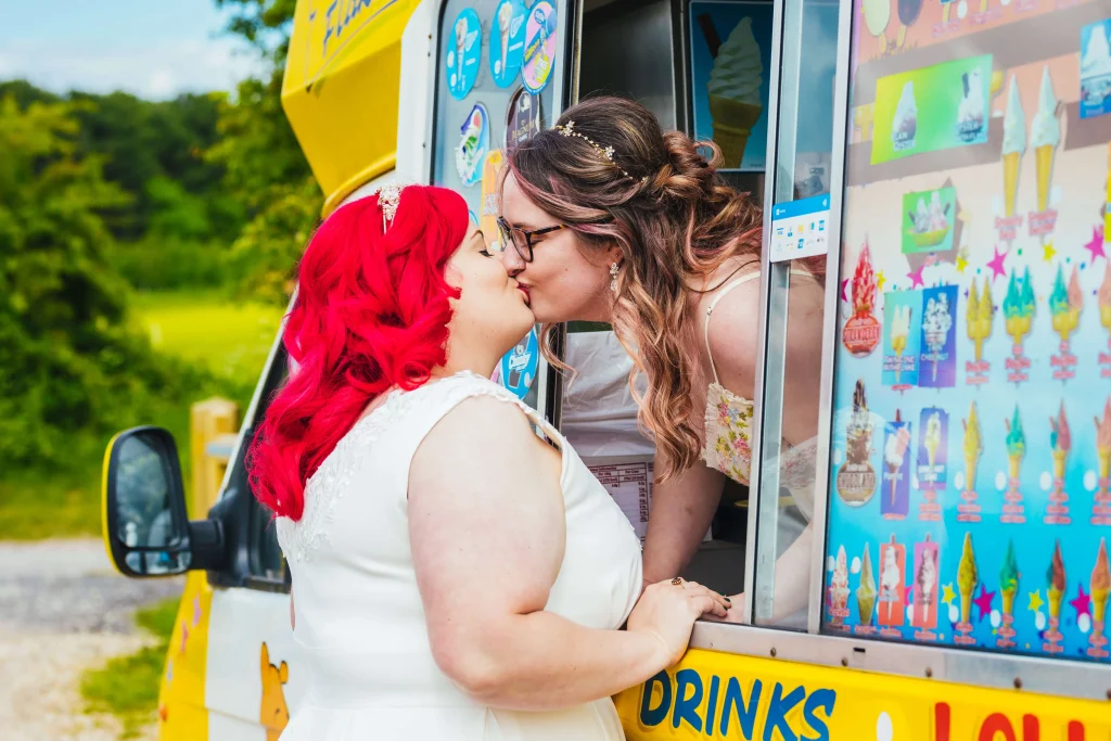 couple on wedding day. One bride is leaning out of a yellow ice cream van window to kiss her new wife who has bright red hair