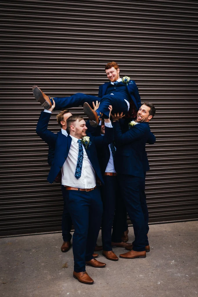 Groom being lifted by his groomsmen in front of a shutter at his wedding venue, they are all wearing blue suits and brown shoes