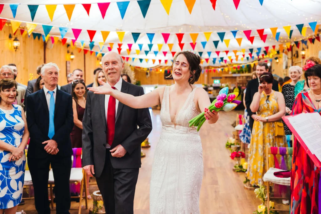Bride walking towards her groom as she reaches the bottom of the isle her hands are out stretched towards him and shes clearly emotional. Her dad and the guests are looking on with pride and emotion.