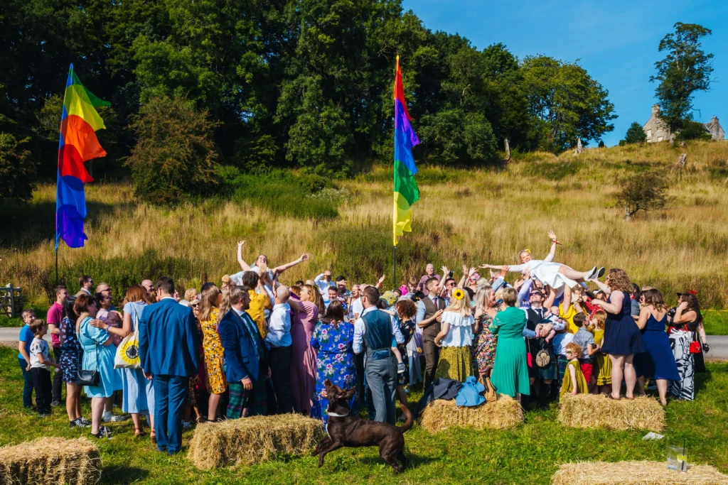 Couple crowd surfing during a full wedding party photo. Their dog can be seen in the forground barking at the drone above as they smile and laugh.