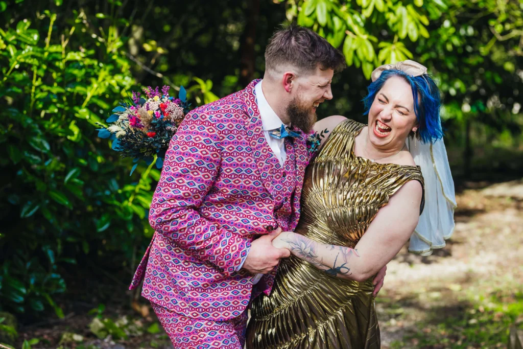Couple laughing together during couples portraits. Groom is wearing the most awesome pink suit whilst the bride has blue hair and wears a gold dress.