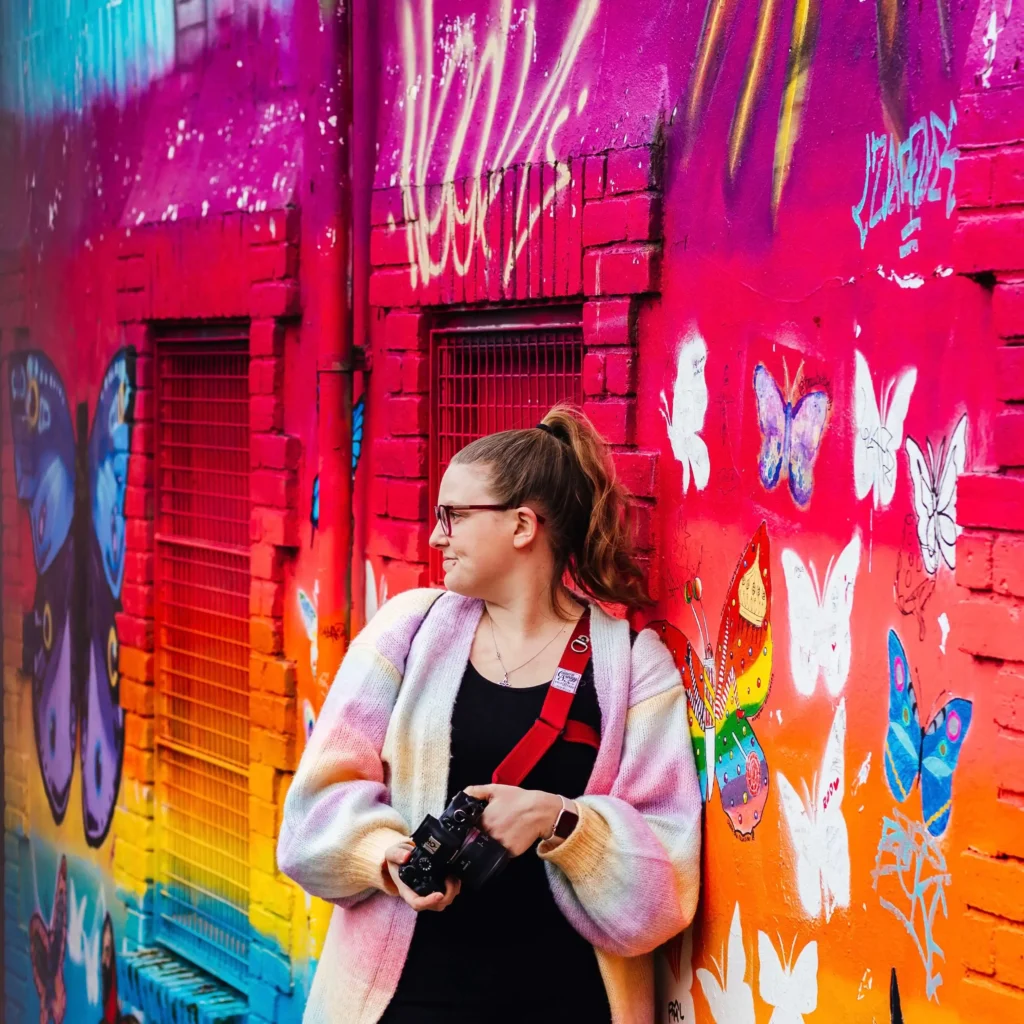 Kirsty stood in front of rainbow graffiti wall with butterflies on it . She is wearing a rainbow cardigan and holding her camera