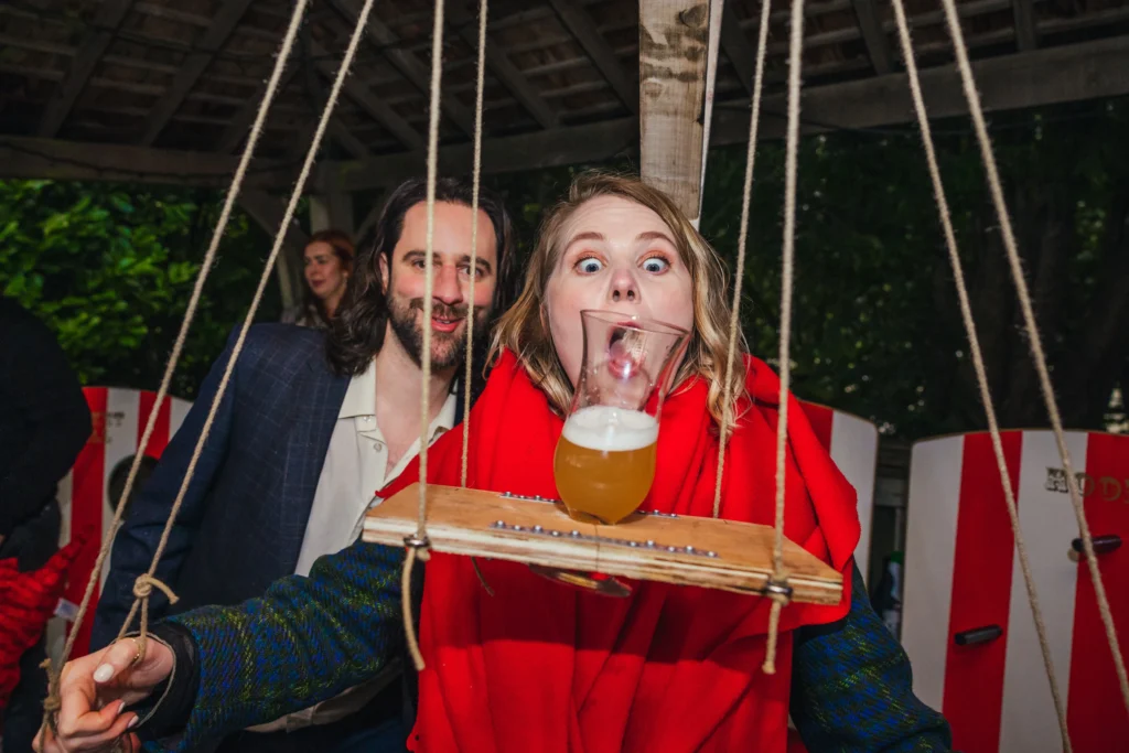 Guest playing beer puppeteer which invloves using strings to bring the glass to your mouth and tipping it so you can drink it without spilling it. The person playing is pulling a wide eyed funny face and the guest behind her is smiling so big.