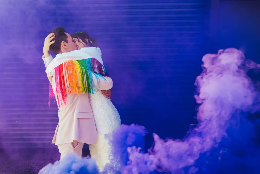 Wedding Photography Nottingham - Wedding couple stood in front of a shutter surrounded by purple smoke. Bride is wearing a thrilly rainbow jacket and is hugging the groom