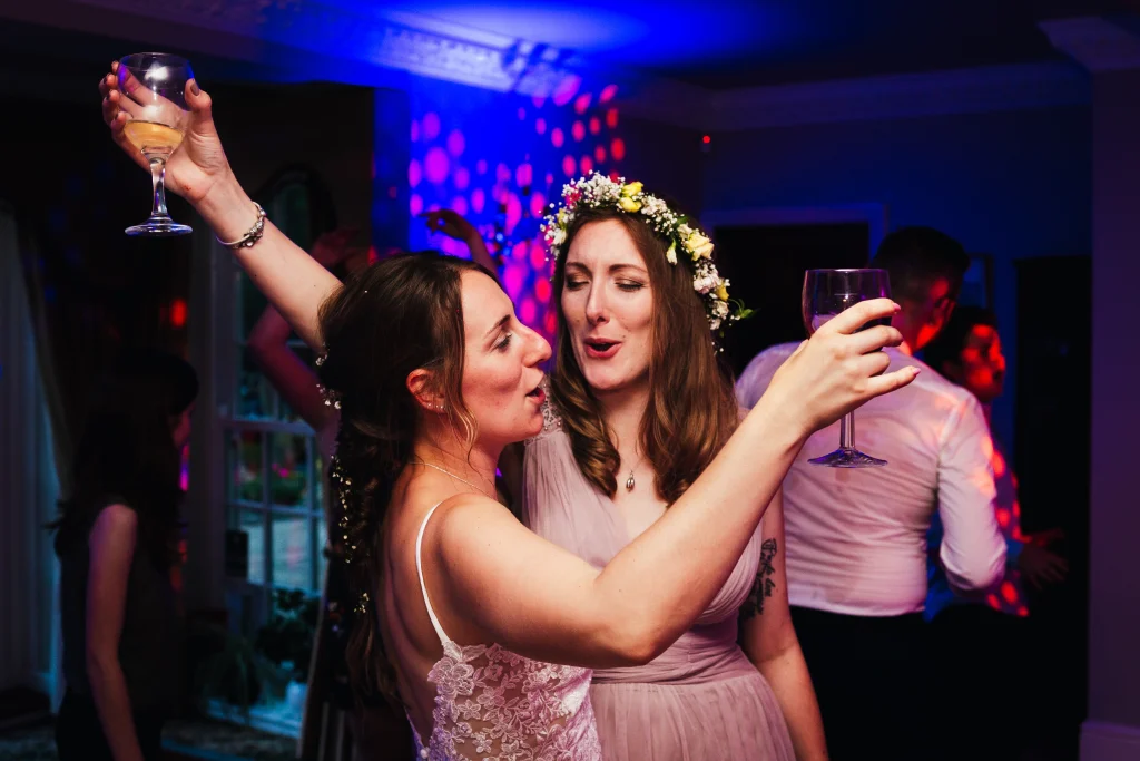 Bride and bridesmaid dancing with their hands in the air, the room behind them is lit by purple lights