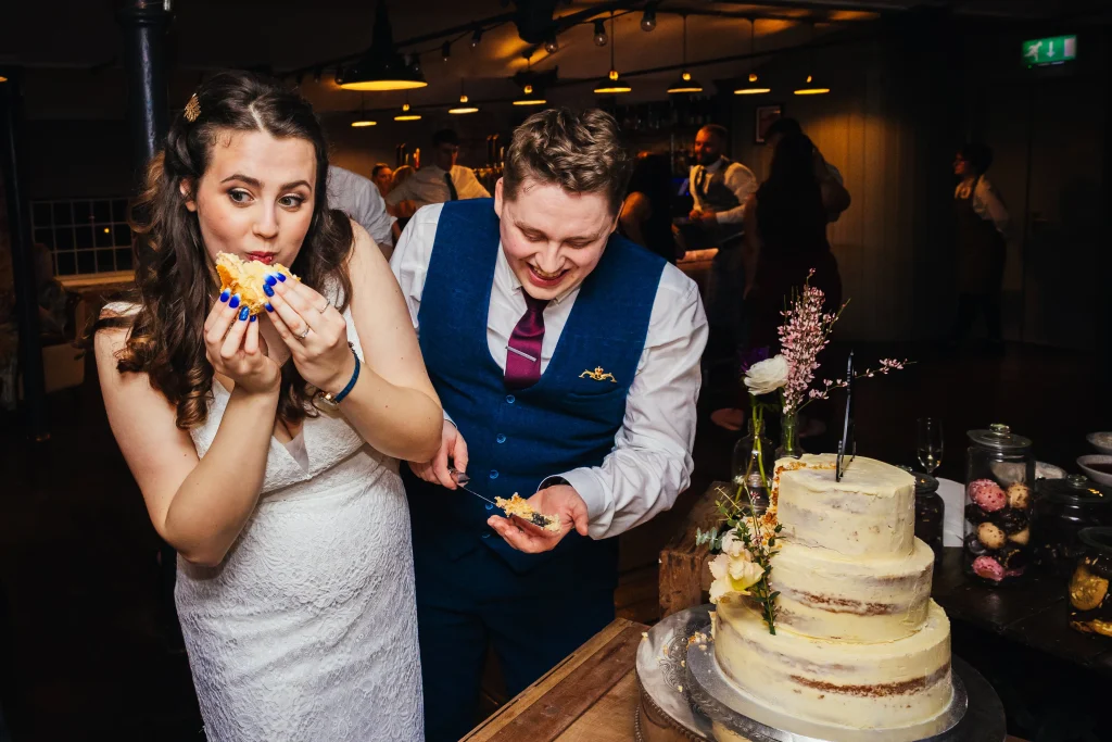 Couple eating cake during their wedding reception - Bride is eating cake whilst groom smiles behind her