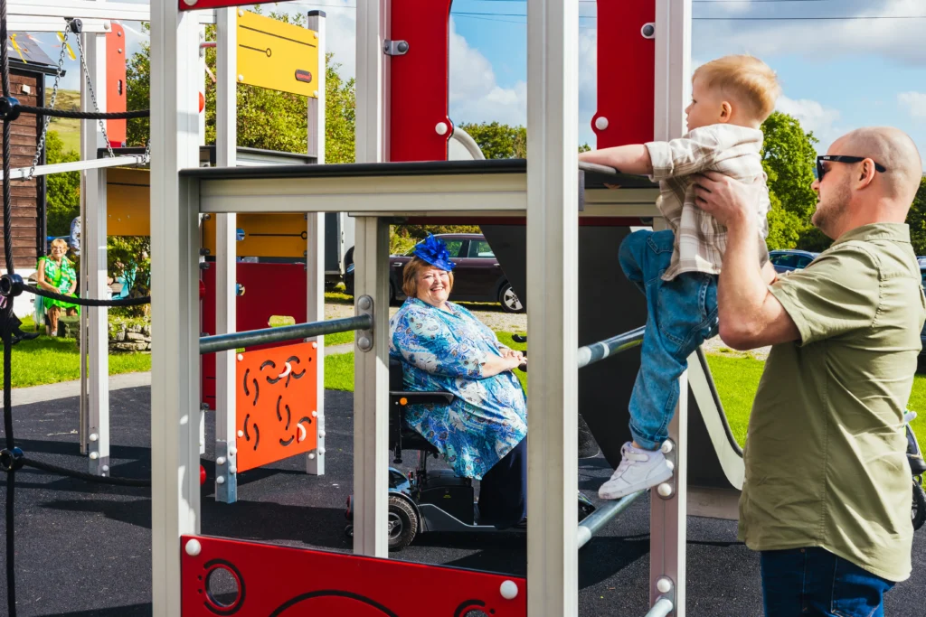 Child climbing on climbing frame being held by dad. Grandma sitting in her scooter watching and is framed by the park