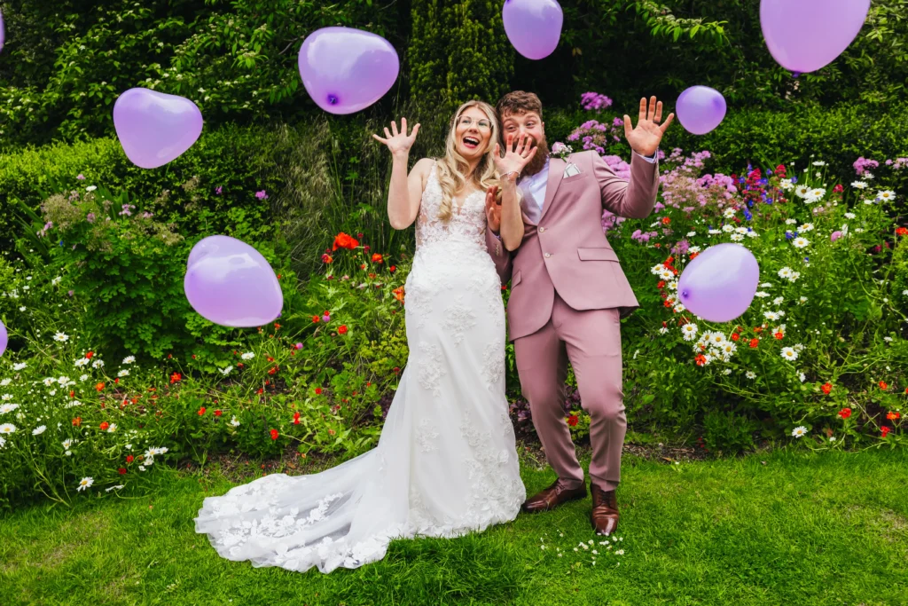 Couple making scared faces as purple baloons are being thrown at them on thier wedding day. They're stood in a beautiful lush green garden. Groom is wearing a pink suit