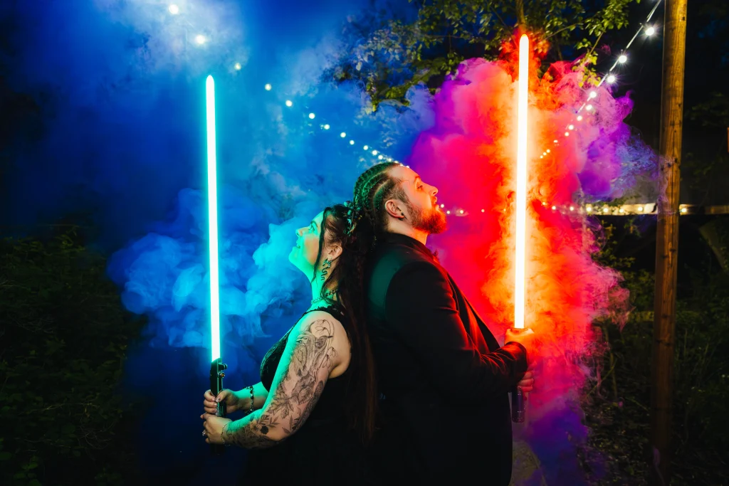 Couple posing with light sabres that are blue and red backed by a purple smoke bomb.