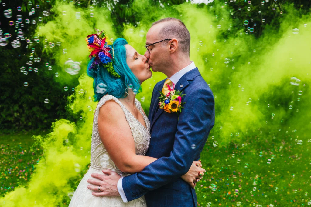Couple stood in front of yellow smoke grenade and bubbles. Bride has bright blue hair and is wearing a flower headdress
