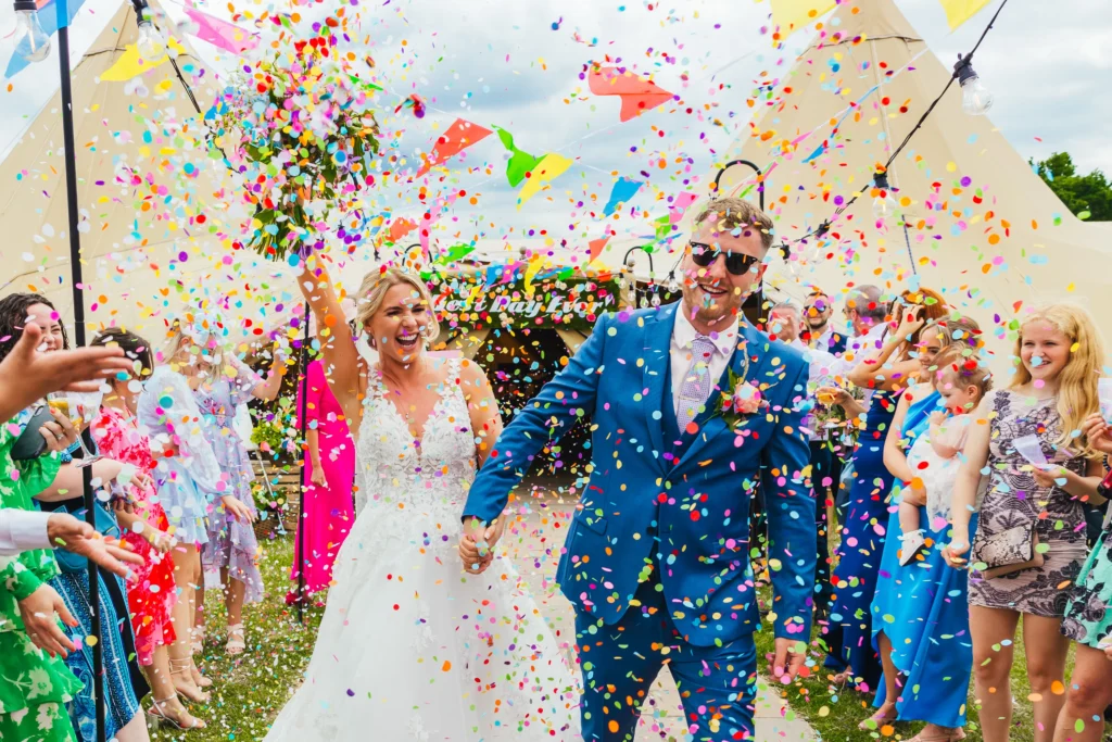 Couple walking through confetti at their tipi wedding the colours are bright and vibrant and all their guests are really enjoying the moment