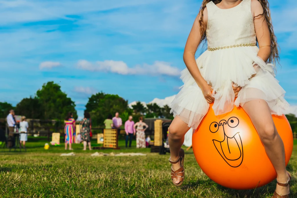 Child on space hopper you can see their face but you can see their bridesmaid dress and sandals. The face on the orange space hopper is the main focus.