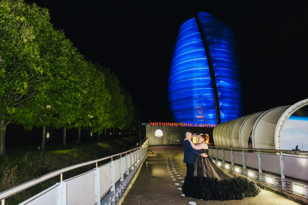 National Space Centre Wedding - Couple posing outside of National Space Centre in the dark it's lit up blue. The bride is wearing a black dress groom is in a suit.