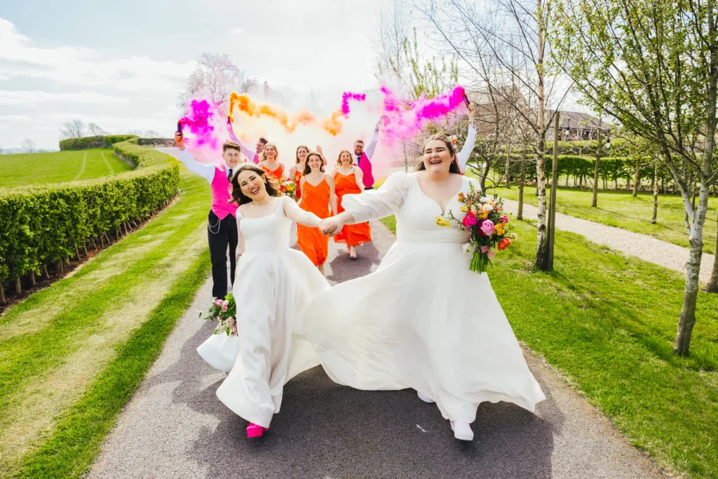 Couple swinging arms walking in front of their wedding party who are dancing and smiling holding colourful smoke. Both brides are holding beautiful colourful bouquets