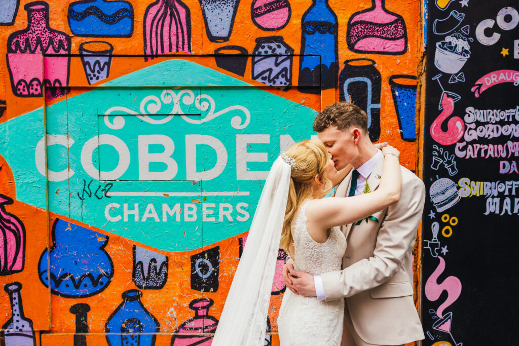 Couple stood in front of bright orange graffiti wall kissing a drinks and food menu can also be seen behind them