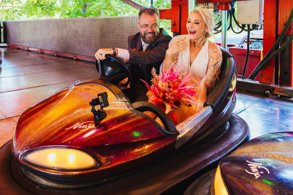 Couple on bumper cars during cocktail hour at their reception bride has a bright pink bouquet on her lap and a massive smile whilst the groom drives they are recording themselves on a go pro
