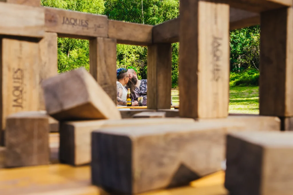 Couple kissing shot through giant Jenga pieces at woodland wedding venue neither know the camera is there.