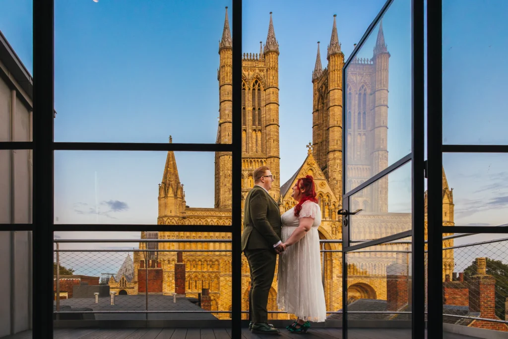 Couple stood on hotel balcony with Lincoln Cathedral in the background. Photographer is stood insideshooting through the open glass door.