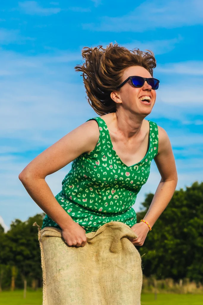 Lady jumping during sack race her hair is flying in the wind and she's laughing. She's wearing a green dress and shades