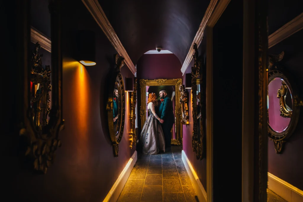 Couple stood in dark purple hallway of mirrors at their wedding venue. The bride wheres a black dress and the groom a green suit. You can see their reflections in the mirrors