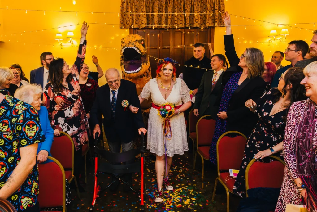 Bride being walked down the isle by future father in law and a dinosaur whilst guests look on in shock, smiling and laughing.