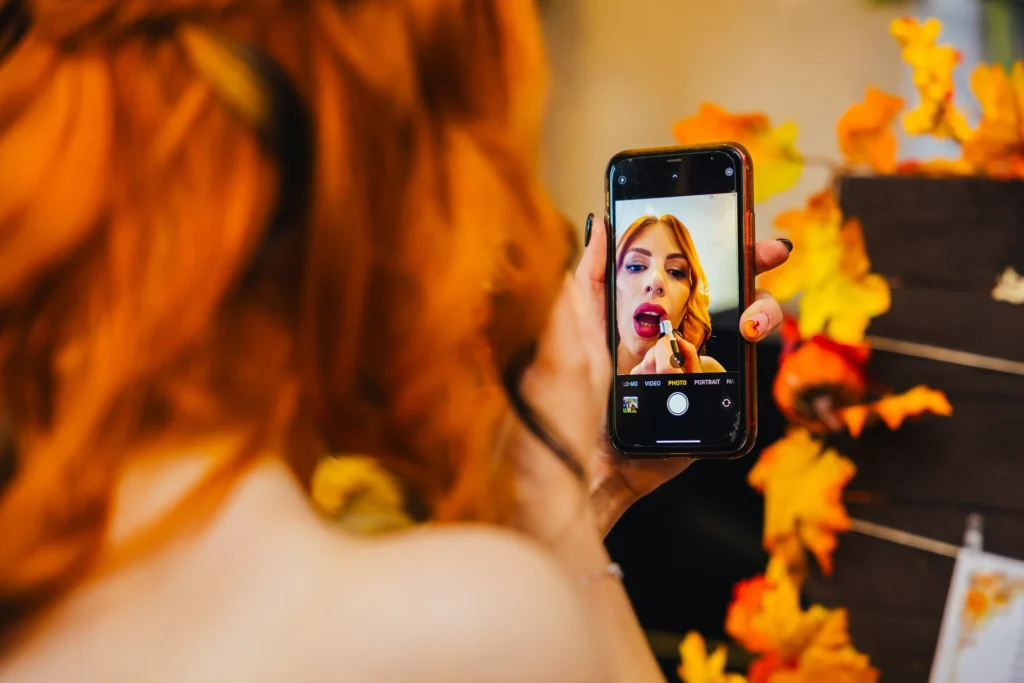 Bride fixining lipstick in her phone after her wedding ceremony her beautiful red hair is perfectly off set with the autumn leaves decorating the venue