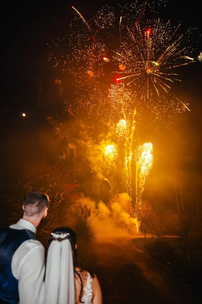 Couple watching fireworks display at the end of their November wedding ceremony the sky is lit up orange and red.