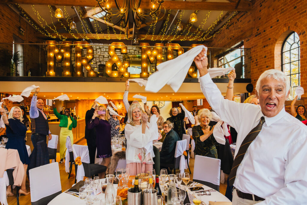 Wedding guests waving their napkins in the air as couple come into their wedding breakfast. Everyones smiling and and some are clapping.