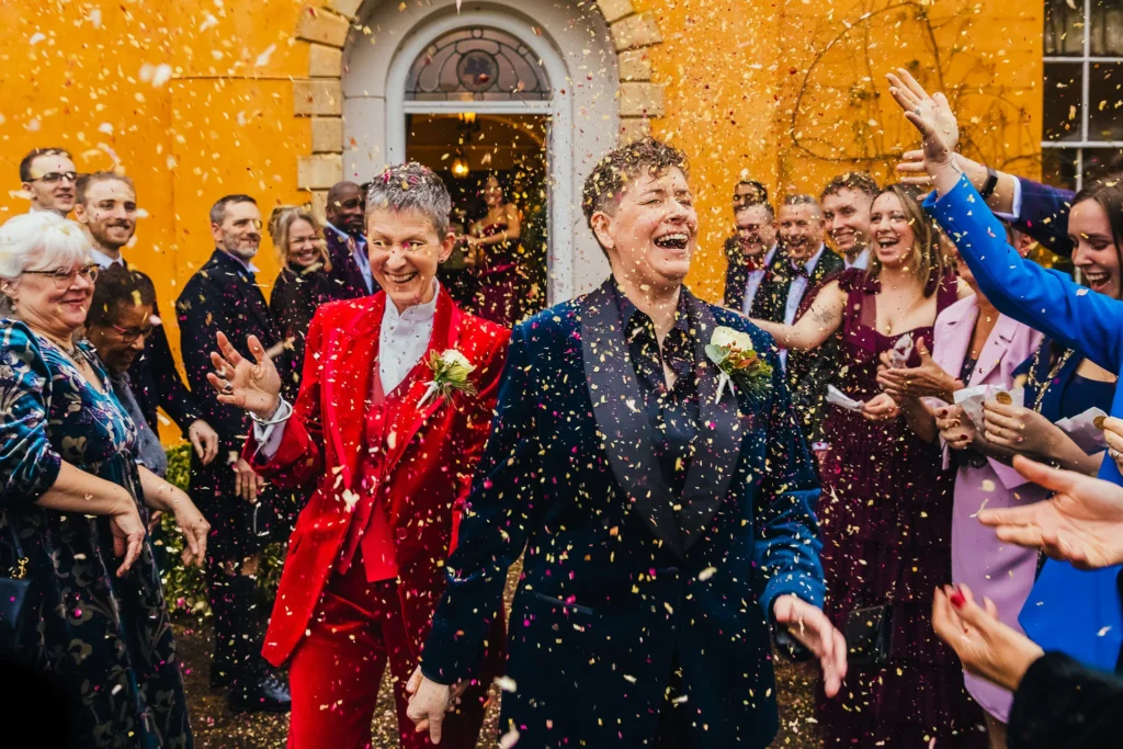 Couple during confetti on their wedding day. One wears a blue suit and the other red both are velvet. Their faces show lots of joy
