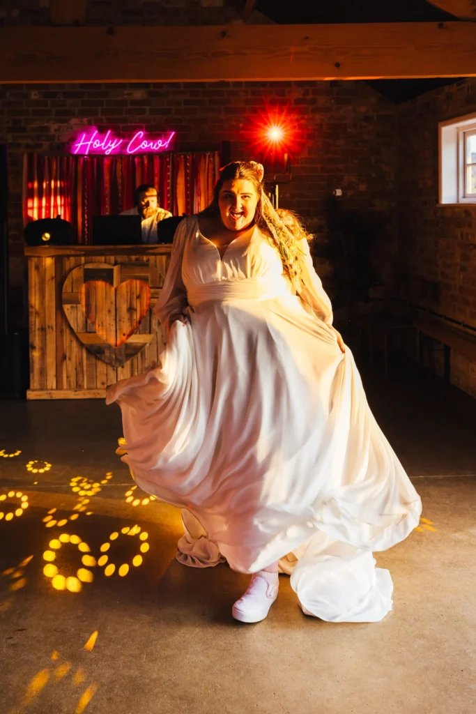Bride dancing and swishing her dress on the dancefloor at their wedding. There's a bright pink sign behind her that says holly cow.