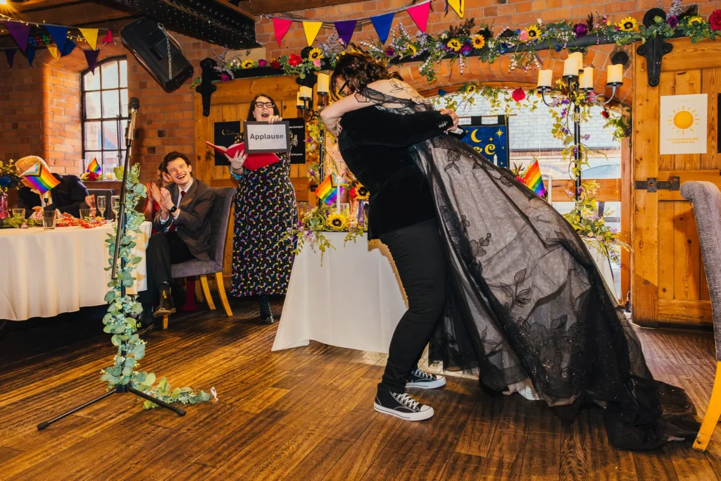 Couple embracing after thier first kiss. One is being lifted of the floor by the other and their celebrant is in the background holding a sign that says applause.