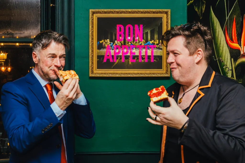Two wedding guests eating pizza stood in front of a sign that says bon appetit during a air bnb wedding reception