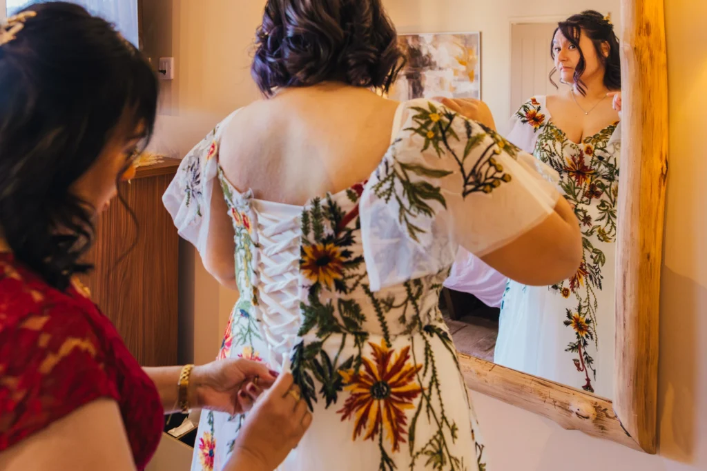 Bride looking at her reflection in the mirror whilst her mum does up her wedding dress. Her dress is white with embroidered flowers on it