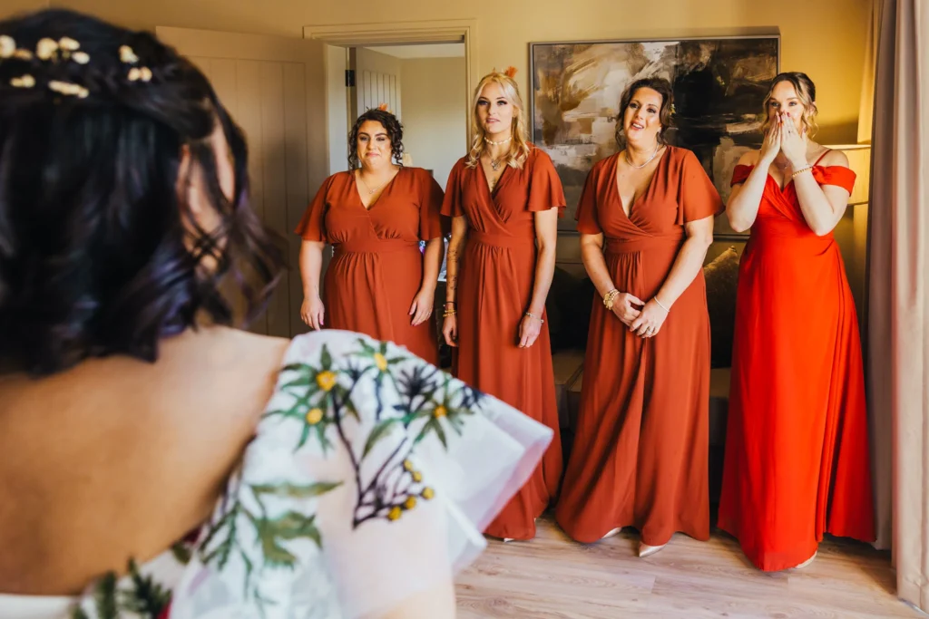 Bridesmaids seeing bride in her wedding dress for the first time all of them are wearing orange dresses and emotional expressions. One has her hands in front of her face.