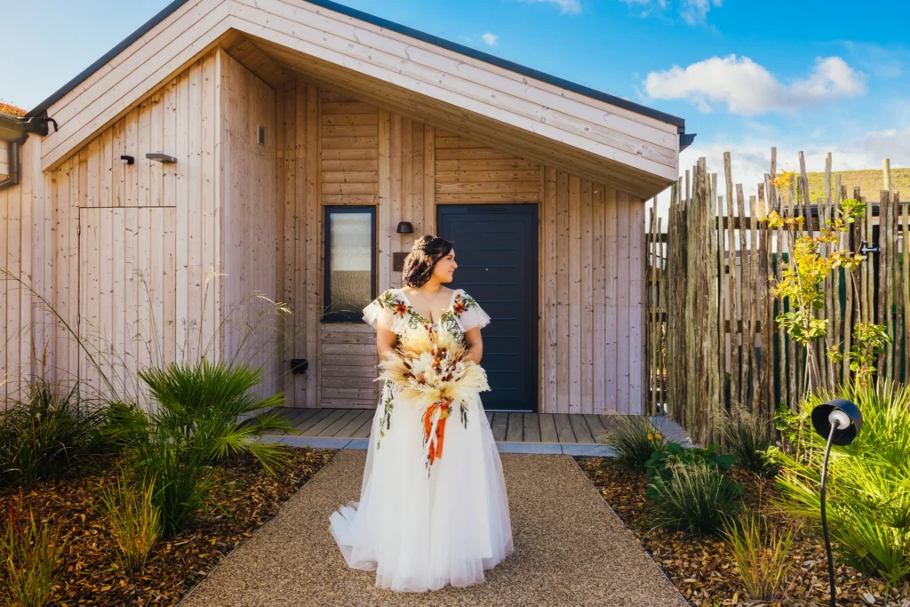 Bride leaving her lodge on the way to her wedding she's holding an autumnal bouquet and her dress is white with embroidered flowers