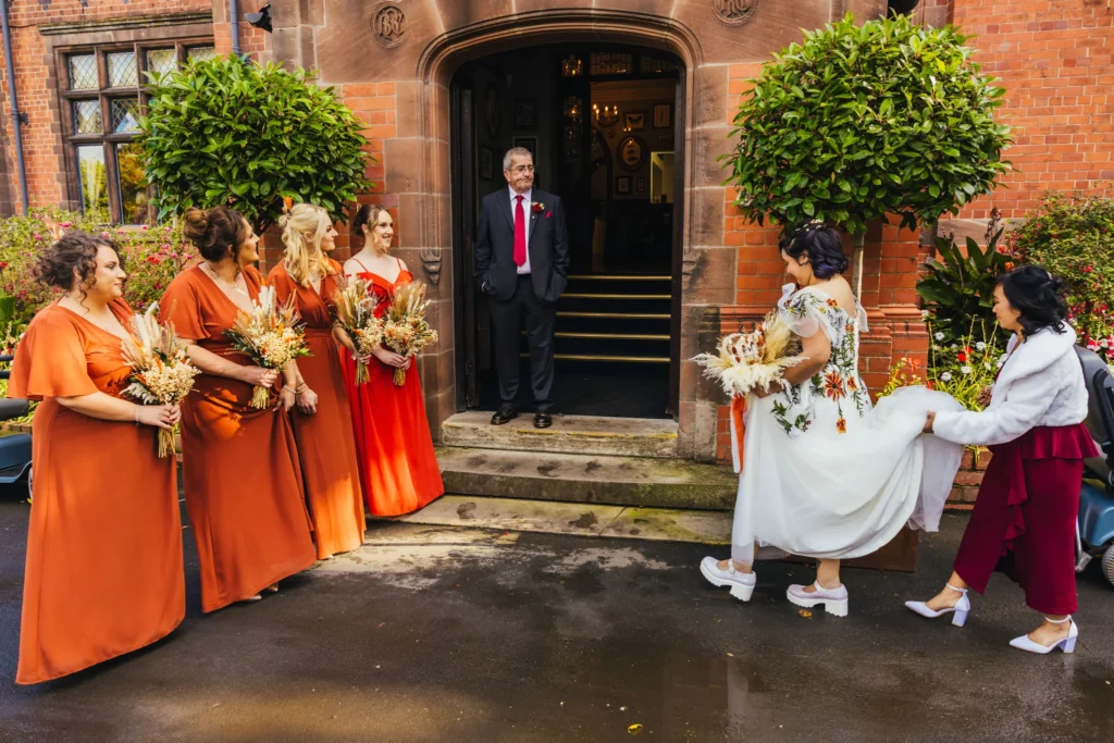 Bride walking to the entrance of The Oakfield at Chester Zoo where her dad is waiting on the front step. Her dad and bridesmaids are waiting for her and her mum is carrying her dress there's lots of autumnal colours