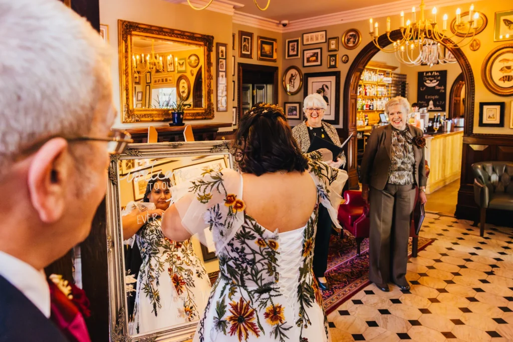 Bride is fixing her dress in a mirror that has welcome to our wedding printed on it just inside the lobby of The Oakfield at Chester Zoo, hee dad is behind her and the registrars are waiting on her smiling
