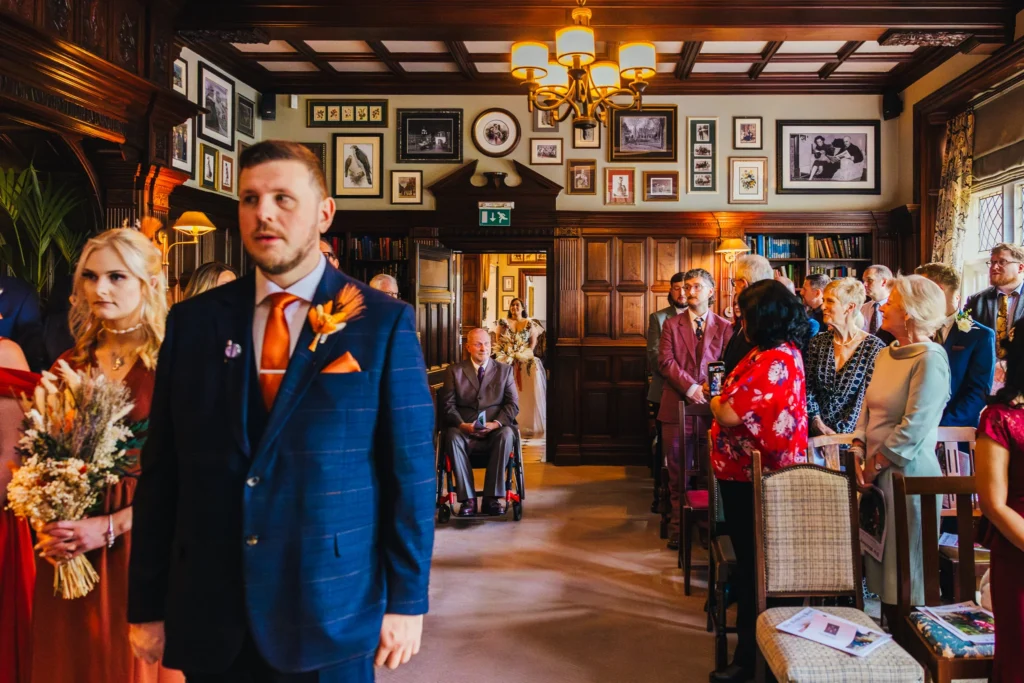 This picture shows the very nervous groom waiting at the top of the isle in a blue suit with an orange tie. all the guests are in the background and you can just see the bride whos about to come through the door in the background