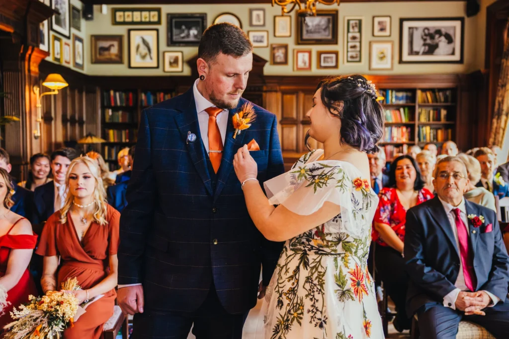 Bride fixes grooms pocket square during the ceremony she wears a white dress with embroidered flowers on it and he wears a blue suit with orange accessories.