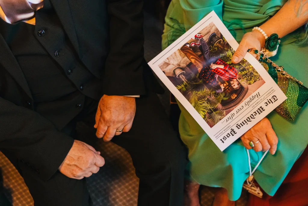 close up of wedding guests one in a suit and one in a green dress holding the couples wedding news paper which has the headline of happily ever after with a picture of them underneath
