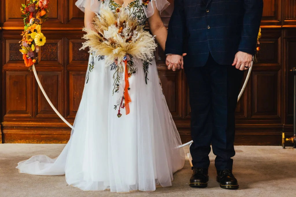 Close up of the bottom half of a bride and groom who are holding hands. Bride also holds her beautiful autumnal bouquet.