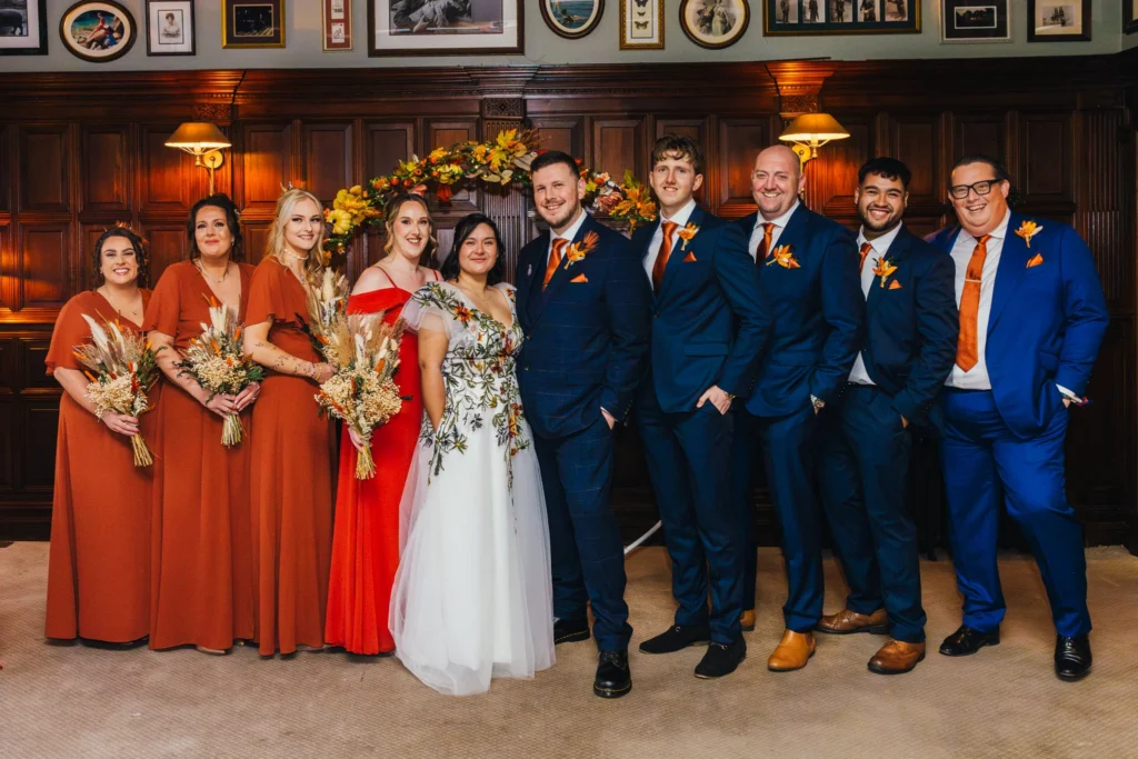 Fun group shot of the whole wedding party. Bridesmaids are in orange groom and groomsman are all in blue with orange accessories. Bride has her hand in the pocket of her dress which has embroidered flowers all over it