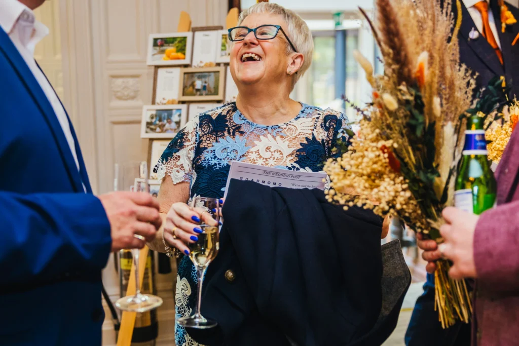 Guest laughing she's holding a glass of fizz and is wearing a blue dress. she is being framed by one of the bridesmaids bouquets