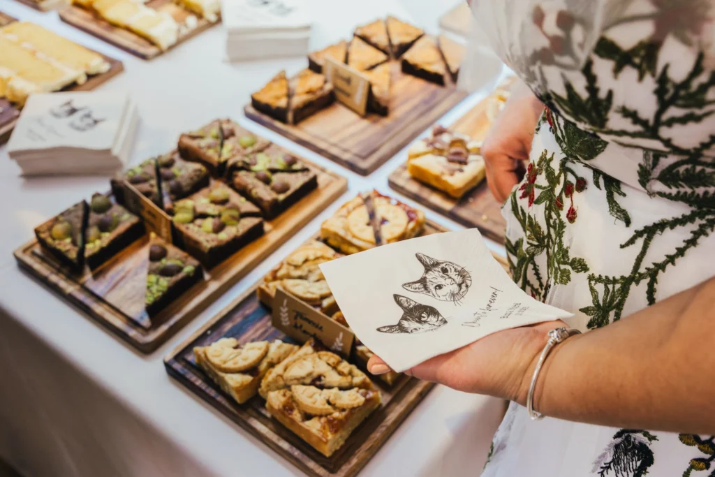 Bride getting some cake during drinks reception the napkin she is holding has a drawing of her cats printed on it. You can see all the yummy cake in the background
