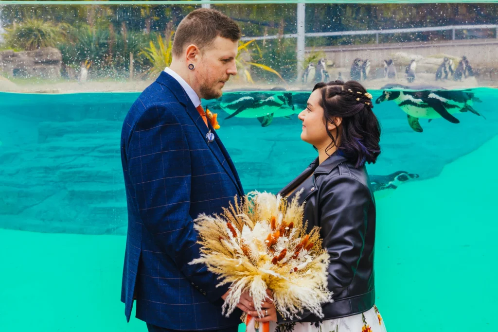 Couple stood in front of penguin tank at Chester zoo. Bride is holding a autumnal bouquet and is wearing a leather jacket groom is in a blue suit.