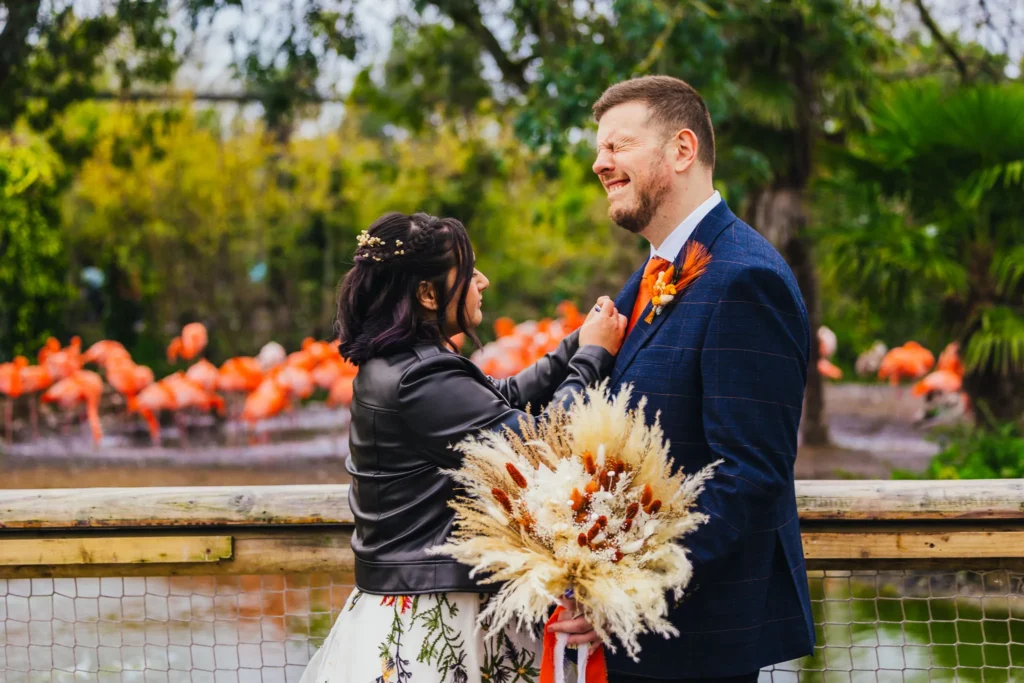 Groom is pulling funny face whilst his bride is sorting his button hole you can see pink flamingos in the background