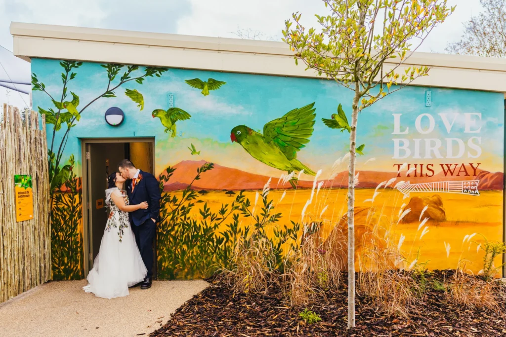 couple stood kissing in the doorway to the love bird enclosure at Chester Zoo. There is wording on the wall that says love birds this way with an arrow pointing directly at them