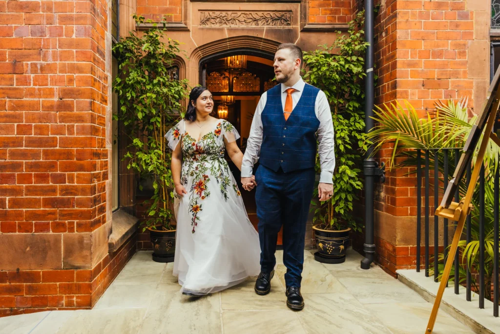 Couple walking into their recpetion holding hands groom haas taken off his jacket and is wearing a blue waistcoat bride is in a white dress with embroidered flowers on it