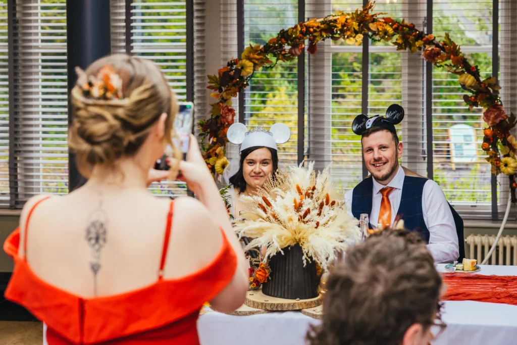 Couple sat at their sweet heart table they're wearing Disney ears whist their maid of honour takes their picture on her phone