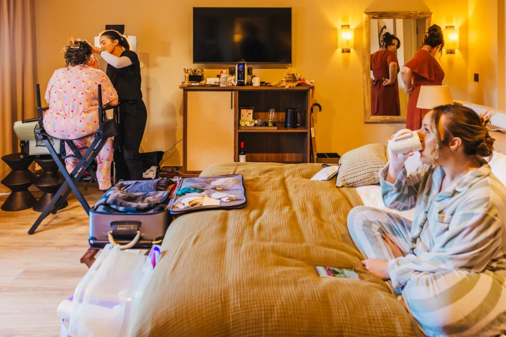 Bridesmaids getting ready in a cabin at The Reserve at Chester Zoo one is in her pjs getting her make up done, another is looking in the mirror doing the bow on her orange dress up and the third one is drinking a coffee on the bed in her pjs.